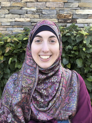 A pale skinned woman facing forward and smiling, wearing a Muslim head covering called a hijab that is brightly colored. She wears a pink long sleeve shirt and stands in front of a stone wall with a green hedge partially covering the wall in the bottom two-thirds of the image.